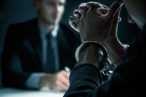 A man in handcuffs waits pensively as a lawyer sits across from him and writes